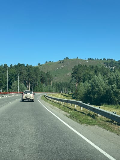 A light-colored Suzuki SUV is driving on a paved road in Souzga, Russia, on a clear, sunny day. The road curves to the right and is bordered by a metal guardrail on the right and a red barrier on the left. On the left side of the road, there are several lampposts. Beyond the road, there are dense green pine trees and lighter green deciduous trees. In the background, a grassy hill with scattered trees rises towards a clear blue sky. The car is the main subject, and its rear is visible as it drives away from the camera. The environment is natural and rural, with evidence of infrastructure for a road.