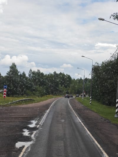 The image shows a wet asphalt road curving to the left, with several puddles present on the road surface. The road is lined with grass and trees on both sides. Several streetlights are visible along the right side of the road. In the distance, there are a few cars and a truck on the road. A traffic sign indicating a turn to the left and a directional sign with red and white chevron markings are visible on the left side of the road. The sky is cloudy. The scene is consistent with a rural or suburban road in Savinskoye Sel'skoye Poseleniye, Russia, likely during or after rainfall.