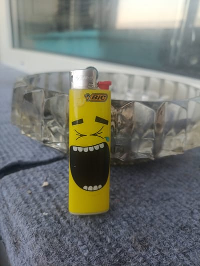 A yellow BIC lighter with a cartoon face is placed on a gray, textured surface. The lighter has a wide-open mouth with teeth and a tear falling from one eye. Behind the lighter, a clear glass ashtray with a faceted design is visible. The background is blurred, showing a window with a light blue tint. The scene appears to be outdoors, on a surface that could be a table or windowsill. The location is Pobedinka, Russia.