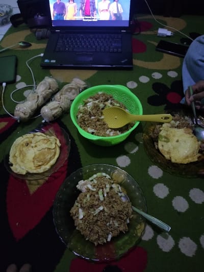 This media file captures an informal indoor scene, likely at night, in Leuwiliang, Indonesia, showing a casual meal arrangement on a patterned rug. A black laptop is prominently displayed at the top, open and showing a video on its screen, featuring several young people. White text, possibly subtitles, is visible on the screen, including the phrase "Kalau pas benci."
Arranged on the rug are several food items. A large green, colander-like bowl holds a substantial amount of fried rice, topped with minced meat and chopped vegetables, and a yellow serving spoon rests inside it. To the right, a clear glass plate holds a portion of similar fried rice, garnished with a fried egg and the same toppings, with a hand holding a spoon and chopsticks eating from it. Another clear glass plate, to the bottom left, also contains fried rice with toppings and a metal spoon. Beside this, a fourth clear glass plate holds several folded, thin, yellowish items resembling omelets or flat pancakes.
Two clear plastic bags, each containing several light-colored, crinkled snacks, possibly crackers or kerupuk, are positioned between the laptop and the food. A dark-colored smartphone is visible, connected to a charger near the laptop, while another phone and a white charger block are seen further right. A black bag or case is situated behind the laptop. The dim lighting and direct placement of items on the floor suggest a relaxed, comfortable, and intimate setting for sharing a meal while enjoying media. At least one person is actively eating, and another person's foot is visible in the bottom left corner, indicating the presence of multiple individuals.