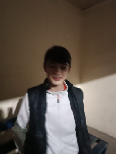 The image features a young male, likely a teenager, captured from the chest up, against a plain, light-colored background. He has short, dark hair and appears to be smiling or mid-speech, though his facial features are somewhat blurred, making a precise expression difficult to ascertain.
He is wearing a white long-sleeved top, possibly a quarter-zip pullover, with a short zipper at the collar. A subtle orange trim is visible on the inside of the collar of the white shirt. Over this, he wears a dark-colored, likely black or dark blue, puffer vest. The lighting in the scene is dim, casting shadows on the left side of the background wall. The background consists of a solid, light-toned wall, appearing to be indoors. There are indistinct shapes or furniture parts visible in the lower corners of the frame, particularly on the left, but they are too blurry to identify clearly. The overall image is blurry, especially the subject's face, suggesting motion blur or an out-of-focus shot. No text is visible in the image.