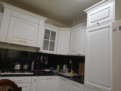 This is a photograph of a clean, well-lit kitchen, likely in Belidzhi, Russia. The cabinets are white with a classic design, featuring ornate detailing on the upper cabinets and simpler flat fronts on the lower ones. The backsplash is made of black marble with white veining, creating a dramatic contrast with the white cabinetry.
On the stovetop, there are two silver kettles and a pot, suggesting that someone may have been cooking or preparing a hot beverage. A collection of knives is neatly arranged on a magnetic strip on the backsplash. Various cleaning supplies and dishes are visible on the countertop near the sink, indicating recent use or ongoing tidying. A wooden cutting board stands upright against the wall.
The overall impression is one of order and functionality. The lighting is bright, suggesting it's daytime, though the specific time cannot be determined. There are no people visible in the image, and the scene is static, offering a snapshot of a domestic space. The focus is on the arrangement of objects and the architectural details of the kitchen. No text is visible in the image.