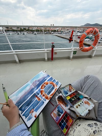 The image depicts an artist sitting on what appears to be the deck of a boat, possibly a ferry, near Aegina, Greece, capturing the scene in a watercolor painting.

The artist is positioned with their legs visible, serving as a surface for a sketchbook and watercolor supplies. A clipboard holds the sketchbook, where a watercolor rendition of a life preserver attached to the boat's railing is in progress. Palettes containing various watercolor paints, along with brushes and potentially a water container, are situated near the sketchbook. The artist's hand is seen holding a brush or pen, actively engaged in the painting process.

In the background, the railing of the boat is visible, and beyond it, a coastal cityscape of Aegina stretches out, featuring a waterfront with buildings, boats, and the sea. A life preserver hangs on the railing, mirroring the subject of the painting. The sky is overcast. The image conveys a sense of travel and artistic creation, with the artist capturing the essence of their surroundings in a watercolor painting while aboard a boat.