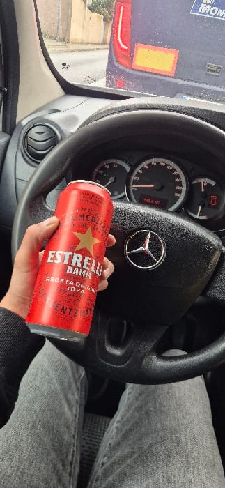 The image shows the interior of a Mercedes-Benz vehicle, from a first-person perspective, within the city of La Garriga, Spain. A person's left hand, clad in a dark sleeve, holds an upright red aluminum can of Estrella Damm beer. The can is marked with a gold star logo, "ESTRELLA DAMM," and "RECETA ORIGINAL DESDE 1876." Below the black steering wheel, which features a silver Mercedes-Benz emblem, the person's lap covered by grey jeans is visible. The vehicle's instrument cluster indicates a speedometer reading of zero, an odometer displaying 290.3 KM, and an illuminated red warning light depicting a car with an open door. Through the moisture-speckled windshield, the rear of a dark-colored van is visible immediately ahead. This van displays a vertical red taillight assembly on its left, a rectangular yellow panel, and a partially discernible white decal. Light-colored building facades are present in the distant background.