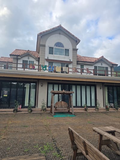 This is a sideways, medium shot of a light-colored, two-story building with a terracotta tile roof. The building has a modern architectural style with several large windows. Decorative elements, such as cutouts of horses and a fruit motif, are visible on the exterior, suggesting a whimsical or perhaps child-friendly establishment.

In front of the building is a rough, brown, mulched area with some sparse green growth. To the right of this area, there are two weathered wooden picnic tables. A wooden swing set with a large wooden reel at its base is positioned near the building's entrance. Potted plants are placed along the building's facade, and a red fire extinguisher is visible near the ground floor.

The sky is overcast with some patches of blue peeking through. The overall lighting suggests it is daytime, likely late morning or afternoon. The scene appears to be an outdoor area of a commercial establishment, possibly a cafe, guesthouse, or restaurant, located in Gumi-si, South Korea.

Visible text on the building includes Korean characters. The larger sign appears to read "신라" (Shilla). Below this, there is smaller text and a fruit graphic with text that appears to say "행복" (Haengbok) and "농장" (Nongjang), suggesting "Happy Farm" or a similar name related to happiness and agriculture. Another smaller sign on the lower right window is illegible. There are no people visible in the image.