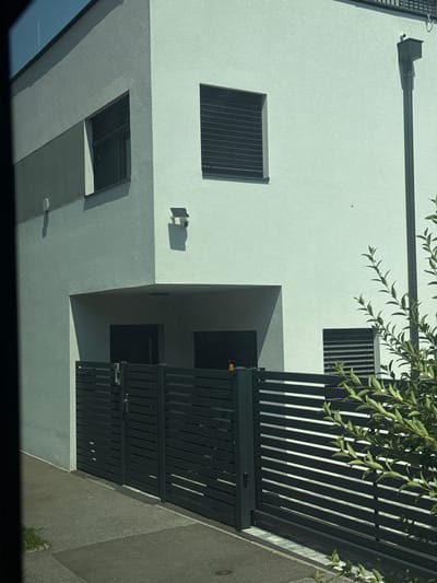 The image shows a modern house with a grey and white facade, a black metal fence, and a concrete sidewalk. The house has several windows with dark louvers. A downspout runs vertically along the right side of the house. There are also two security cameras visible on the exterior of the building. The scene is captured during the daytime, with clear sunlight casting shadows. The house is situated in the city of Wolfsthal, Austria, as indicated by the context. The environment appears to be a residential area with greenery visible in the foreground.