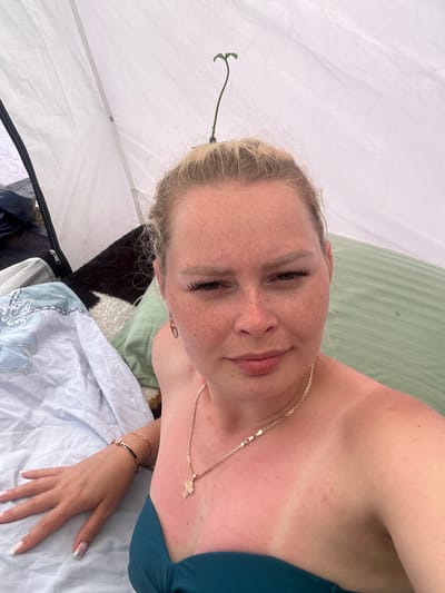 The image shows a close-up, selfie-style shot of a woman with blonde hair and freckles, who appears to be inside a tent. A small green sprout is growing from what appears to be a clip in her hair. She is wearing a teal-colored top and a gold necklace. Her skin shows signs of sunburn on her chest and shoulders. To her left, a light-colored blanket with a blue and white pattern is visible, along with other bedding. The background is primarily the white interior of the tent. There is no visual information within the image to suggest the location is the city of Колодезный, Russia.