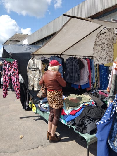 In Chudniv, Ukraine, an outdoor market stall is set up under a canopy. A woman, wearing a brown leather jacket, a red baseball cap, leopard-print skirt, and brown leggings, stands examining clothing. The stall displays a variety of garments, including shirts, pants, and a patterned dress, on hangers and piled on a table. The ground is paved, and the sky above is blue with white clouds.