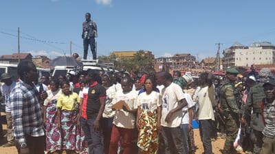 A large crowd gathers on a sunny day. Many wear patterned skirts and t-shirts with printed images. A man in a plaid shirt appears to be speaking. A photographer stands atop a white vehicle, capturing the scene. Military personnel in camouflage uniforms are present, seemingly providing security. Buildings and houses form the backdrop, indicating an urban or semi-urban setting.