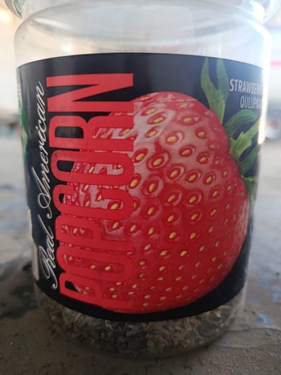 A close-up, slightly angled shot shows a clear glass jar with a label featuring a large, red strawberry illustration and bold red text that reads "Real American POPCORN". The text is stylized and appears to be embossed. To the right of the strawberry, the word "STRAWBERRY" and "QULUPNA" are visible in smaller white text. The jar contains a dry, granular substance at the bottom, appearing to be popcorn kernels or dried herbs. The jar sits on a rough, textured surface, possibly concrete or stone. The background is out of focus, but hints of a blurry, outdoor environment suggest a bright day. The image's focal point is the product label on the jar. There are no people present in the image. The scene does not provide any specific indicators of its location within the city of Karshi, Uzbekistan.