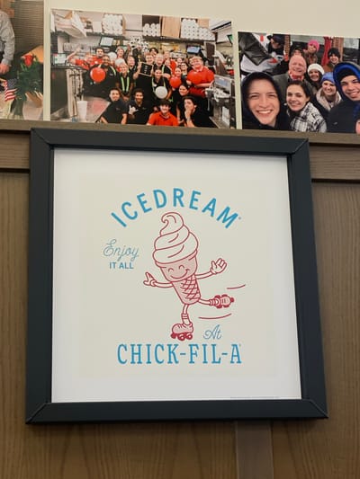 The image shows a framed print with an illustration of an anthropomorphic ice cream cone wearing roller skates, with the text "ICEDREAM ENJOY IT ALL AT CHICK-FIL-A" printed on it. The print is hanging on a wooden wall. In the background, there are several photographs of groups of people. One photograph shows a group of Chick-fil-A employees behind a counter. Another photograph shows a group of people posing for a picture outdoors. The location is identified as Hagerstown, United States of America.