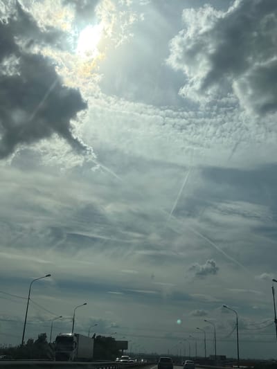 This image captures a daytime highway scene in Razmetelevo, Russia, observed from a vehicle. The sky is largely covered by a diverse array of clouds, with the sun prominently visible in the upper-left quadrant, partially obscured by dense, dark gray cumulus-like clouds. Bright sun rays emanate from behind these clouds, illuminating lighter, more fragmented altocumulus and stratocumulus cloud formations that stretch across the rest of the sky. Multiple distinct aircraft contrails are visible as thin white lines traversing the cloud cover in various directions, particularly in the upper and middle portions of the frame.
The lower third of the image shows a multi-lane highway receding into the distance. A dark metal guardrail is present in the immediate foreground. On the left side of the highway, a white tractor-trailer truck is traveling away from the viewer. Numerous passenger cars are also visible on the highway lanes, extending into the background. Tall, dark streetlights with curved arms are systematically spaced along both sides of the road. Above the streetlights on the left, utility poles and power lines are discernible. Sparse dark foliage (trees and bushes) lines the highway shoulders. The overall lighting is backlit, causing the foreground elements, including vehicles and infrastructure, to appear in silhouette or darker tones against the bright sky. A circular lens flare or reflection is present in the lower-mid portion of the image.