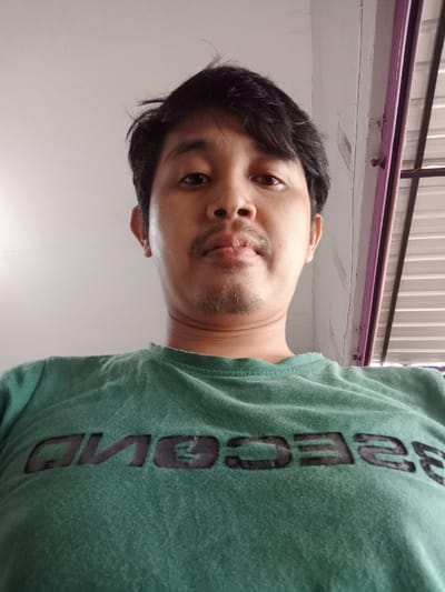 A medium shot displays a man looking directly at the camera from a low angle. He has dark hair, a mustache, and wears a green t-shirt with the word "SECOND" printed in black letters. The background reveals a white wall and a glimpse of what appears to be a building exterior, possibly a metallic roof or paneling, accented with a purple frame.