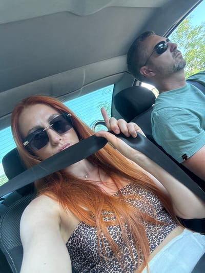 The media file shows two people, a man and a woman, in the interior of a car. The man, on the left, is wearing a light blue t-shirt and sunglasses. He has short, dark hair and a beard. The woman, on the right, has long, auburn hair and is wearing sunglasses and a leopard-print top. Both individuals are wearing seatbelts. The image is taken from a low angle, looking upwards, with the car's interior visible, including the seats and parts of the dashboard. Through the car windows, trees and a bright sky can be seen, suggesting it is daytime. The scene appears to be a candid shot taken inside a moving vehicle. The lighting suggests it is a sunny day. There is no visible text in the media file. The provided location context is Medinski Krš, Montenegro.
