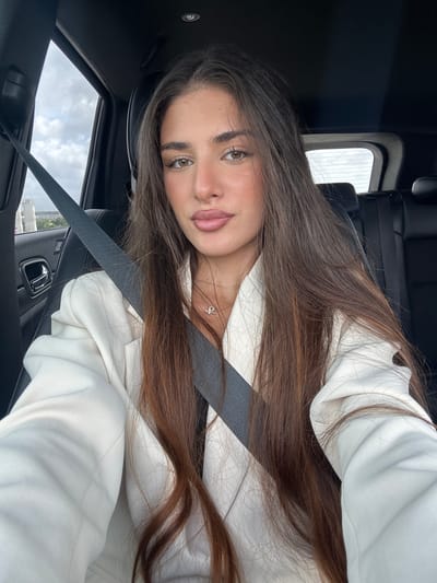A woman is sitting in the driver's seat of a vehicle. She is wearing a white blazer and a seatbelt. The scene is in North Miami Beach, Florida, United States of America. The sky is visible through the sunroof.