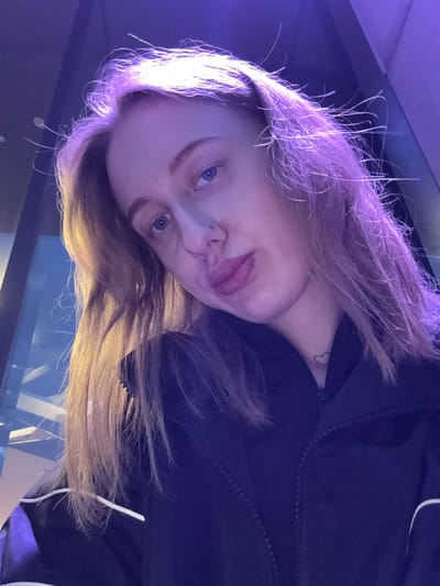 A portrait of a fair-skinned person with light blue eyes and light brown hair under violet-toned lighting. The person has a nose piercing on the left side and a heart tattoo on their neck. They are wearing a dark, possibly black, zippered jacket. The background appears to be a combination of dark and light blue areas, possibly indicating an indoor setting with some reflective surfaces. The image is taken from a low angle, looking upwards towards the person. The city and location are not discernible from the image.