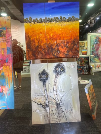 The image shows an art exhibition at the "Dialogue Art" gallery in Surfers Paradise, Australia. In the foreground, a large painting of an emu with distinctive yellow eyes is displayed. To the left, another abstract painting with vibrant colors is visible. Behind these, a panoramic landscape painting of an Australian outback scene with a bright blue sky dominates the upper portion of the image. In the background, more artwork and a person browsing are visible. The exhibition appears to be in a well-lit indoor space with a dark carpeted floor.