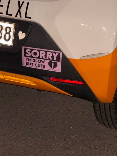 The image depicts a portion of a vehicle, likely a car, parked on a paved surface. The visible section of the car is located near the front driver's side wheel well. The car has a white and orange paint scheme with a black accent. A pink sticker is affixed to the black section, reading "SORRY I'M SLOW BUT CUTE" with a heart and question mark graphic. A silver heart-shaped sticker is visible near the license plate, which partially reads "XL 8". A small vertical red reflector is also present. The tire of the vehicle is partially visible at the top of the frame, indicating the car is parked on a road or parking space. The location is given as Borough of Spelthorne, United Kingdom but there are no aspects of the image which indicate this is the exact location.