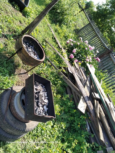 In Nizhniye Vyazovye, Russia, on June 15, 2026, at 13:22, a backyard scene is captured. The foreground features a stack of old tires supporting a metal container filled with charcoal. To the left, a rusty drum, possibly from a washing machine, also contains charcoal and sits on metal legs. A wooden fence made of weathered planks runs diagonally through the background. Adjacent to the fence are several scattered pieces of wood. A cluster of pink peonies and other green foliage with small yellow flowers surrounds the objects, indicating a garden setting. The overall impression is one of a rural or semi-rural outdoor space, possibly prepared for a barbecue.