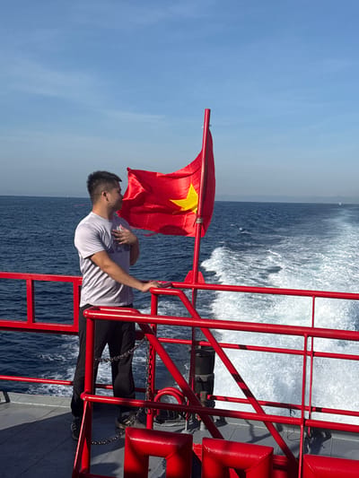 A man stands on the deck of a boat, his right hand placed over his chest, looking towards the Vietnamese flag which is flying on a red pole. The flag is red with a yellow star in the center and is waving in the wind. The boat is moving, creating a wake in the blue ocean. The sky is clear and blue. The scene appears to be set in the city of Dong Son, Vietnam.