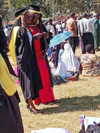 A group gathers outdoors on a grassy field. Several people stand, including two women in graduation gowns and caps. One wears a red dress underneath. They look at a mobile phone. Others are seated, some under a blue and white umbrella. The crowd includes men in casual wear and women in various outfits. A car is visible in the background behind the standing people.