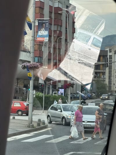 This image captures a street scene in Konjic, Bosnia and Herzegovina. The primary subjects are pedestrians crossing a street, including two women and a child. One woman, wearing a pink top and a patterned skirt, is walking with a white bag, while another woman, dressed in a patterned dress, is holding the hand of a young child wearing pink shorts. Several vehicles are visible, including a silver Volkswagen Golf and a red car. The scene appears to be taking place during the daytime, under what seems to be overcast or slightly hazy weather conditions, as indicated by the lighting and reflections on the car windows.
Notable details include a poster on a building advertising a concert by "DŽENAN LONČAREVIĆ" on "KONJIC 4.7.2016". A flag, likely the flag of Bosnia and Herzegovina, is also visible, with its distinctive yellow stars and blue stripe on a white background. The architecture of the buildings suggests an urban or town setting. The reflections on the car windows are prominent, creating a layered effect that obscures some of the scene. The overall atmosphere seems to be that of a typical day in a European town.