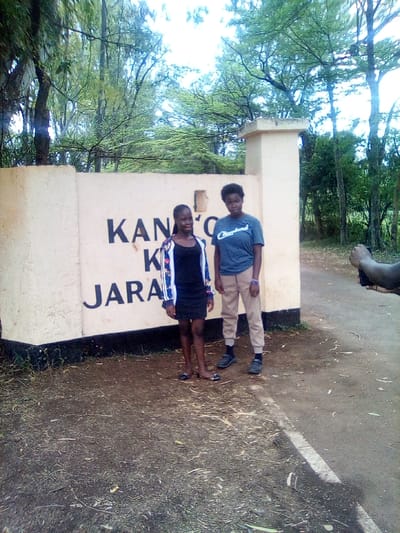 The image displays two young individuals posing outdoors in front of a light-colored concrete wall featuring partially visible black text that reads "KANTO K JARAR." An adjacent light-colored pillar stands to the right of the wall. This scene is located in West Sakwa ward, Kenya.
The person on the left, appearing young and dark-skinned with dark hair, stands facing the camera, smiling slightly. She wears a black sleeveless top or dress under an open, long-sleeved jacket with a white and blue pattern, and light-colored flat shoes. The person on the right, also appearing young and dark-skinned with short dark curly hair, stands slightly behind the first individual, facing the camera with a neutral expression. She wears a dark blue t-shirt with "Cleveland" printed in white script, light brown or beige jogger-style trousers, dark socks, and dark athletic shoes. A blue wristband is visible on her right wrist.
To the far right of the frame, a dark-skinned arm and hand are visible, holding a black smartphone, indicating a third individual is actively capturing the photograph. The ground beneath the subjects is composed of bare earth with scattered dry vegetation. To the right, a path or road surface is visible, featuring a partial white painted line. The background is dominated by a dense canopy of various green-leafed trees under an overcast or light sky.