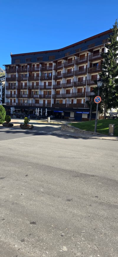 The image shows a street scene in Sallent de Gállego, Spain, on a clear, sunny day. The focal point is a large, curved, multi-story building with white walls and numerous balconies featuring wooden railings and shutters. The building has a dark blue roofline. In front of the building, there are shops and outdoor seating areas for cafes or restaurants, with umbrellas providing shade. To the right of the building, a large pine tree stands tall against the bright blue sky. A street lamp with a "no parking" sign is visible near the tree. The foreground consists of a paved road and sidewalk. There are no people clearly visible in the image, and the scene appears peaceful and unoccupied.