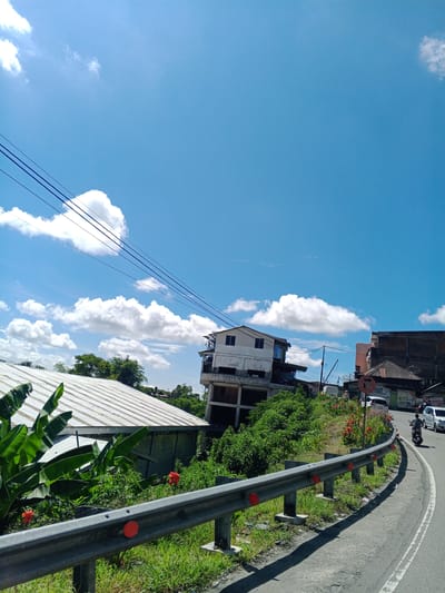 The image depicts a bright, sunny day with a clear blue sky dotted with fluffy white clouds. In the foreground, a paved road curves to the right, bordered by a silver metal guardrail with red reflective markers. Green grass and some scattered debris are visible along the roadside.
On the left side of the road, a white-roofed structure with large green banana leaves in front of it is partially visible. Further back, a multi-story building with white and gray facades stands on a hillside, appearing to be under construction or in disrepair, with visible concrete structures and balconies. Lush green vegetation covers the hillside around the building.
On the right side of the road, a few vehicles are present, including a motorcycle with a rider and at least two cars. More buildings, some appearing to be shops or houses, are visible further up the hill and along the road. A sign with a red arrow indicating a turn is also visible.
The overall scene suggests a residential or semi-urban area in a tropical climate, likely located in the city of Babussalam, Indonesia, given the presence of tropical vegetation and building styles. The presence of power lines strung across the sky further indicates an inhabited area.