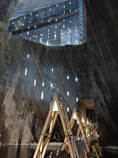 The image depicts the interior of the Salina Turda salt mine in Turda, Romania, a former salt extraction site transformed into an underground amusement park and museum. The scene is characterized by vast, cavernous walls of salt, displaying distinct layered striations in shades of gray and white. Numerous vertical, rectangular lights are embedded at intervals along the walls, casting a cool illumination.
In the foreground, a wooden walkway with a railing curves into the distance, supported by a series of A-frame wooden structures. These structures are also illuminated by triangular white lights at their apexes. The walkway appears to lead over a dark, still body of water at the base of the cavern.
Above, a long, suspended walkway or bridge with wooden planks and metal supports is visible, also lined with lights. A person wearing a red shirt and dark shorts can be seen walking on the lower wooden walkway. The overall impression is of a massive, artificially lit underground space with a blend of natural geological formations and man-made structures for tourism.
