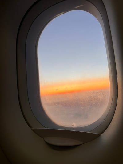 An airplane window frames a view of the sky at sunrise or sunset. The sky transitions from a light blue at the top to a vibrant orange band near the horizon, and then to a hazy, pinkish-grey layer below, possibly clouds or the earth obscured by atmospheric conditions. The window frame, made of a light-colored material, is visible in the foreground, casting a shadow onto the sill. The glass of the window appears slightly frosted or textured. The scene is devoid of people or any discernible urban landscape.