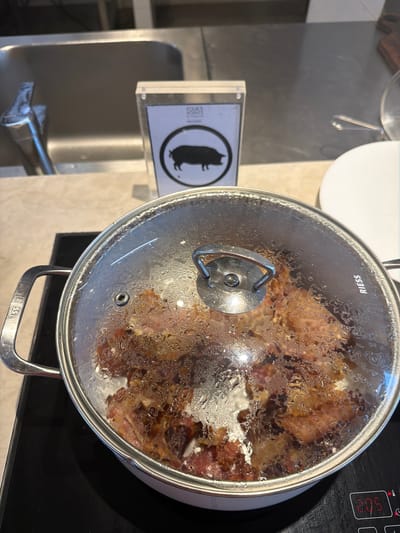 A top-down shot depicts a kitchen setting. A stainless steel pot with a clear glass lid rests on a black stovetop, its contents visible through the condensation on the lid. The pot has silver handles and the word "Riess" imprinted on its rim. The stovetop displays a digital timer reading "3:05". Next to the sink, a sign shows the silhouette of a pig in a black circle, with "FOUR POINTS" printed in small letters. The sink is stainless steel and equipped with a silver faucet. The setting is indoors, likely in a kitchen or dining area of a Four Points hotel in Commune de Diamniadio, Senegal.
