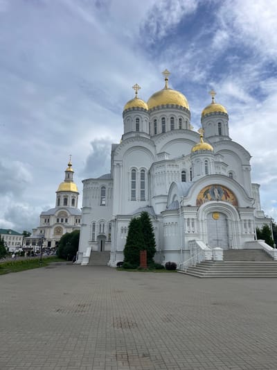 In the city of Diveyevo, Russia, a large, white, multi-domed Orthodox church stands prominently under a cloudy sky. The main structure features numerous golden domes topped with crosses, and decorative golden mosaics above arched entrances. Stairs lead up to the main entrance, flanked by white railings. To the left of the main church, a smaller, taller church with a golden dome and cross is visible, also featuring white architecture and decorative elements. A paved walkway extends in front of the churches, with scattered people in the distance walking on grass and near the buildings. Several trees, including evergreen varieties, are situated around the church grounds. The overall scene depicts a significant religious complex.