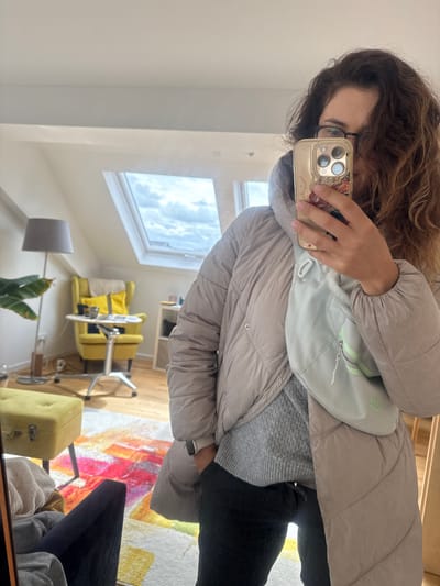 The image shows a person in a gray puffer jacket and gray sweater, with a light blue Nike fanny pack. They are holding a gold iPhone in front of their face, presumably taking a selfie. The person has curly brown hair and is wearing glasses.

The background includes a room with a skylight and a yellow chair. A floor lamp and a plant are also visible. The rug is brightly colored. The image is likely a reflection in a mirror.

It is not possible to determine the location as Ampfing, Germany from the image.