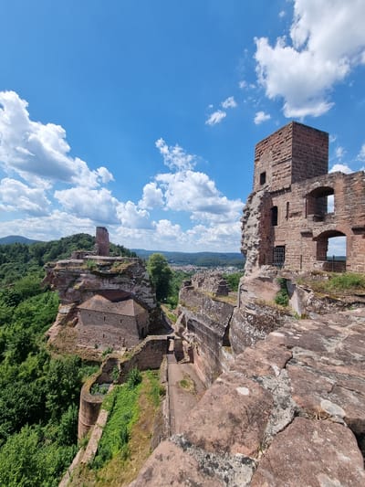 The image shows a medieval castle ruin perched atop a rocky outcrop, overlooking a forested valley and a town in the distance. The castle is constructed from red sandstone and appears to be largely ruined, with crumbling walls, intact towers, and arched windows. The sky is a bright blue with scattered white clouds. The castle is identified as the "Alt-Dahn" castle in Dahn, Germany. The environment is characterized by rugged terrain, lush green vegetation, and a clear sky. There are no people or animals visible in the image. The scene depicts a historical landmark set in a natural landscape.