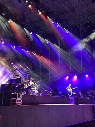 The image depicts an outdoor concert at night in Puerto De Sancti Petri, Spain. A stage is set up with numerous lights casting beams of red and purple light, some of which are partially obscured by smoke. On the stage, two musicians are visible. In the foreground, to the left, a drummer is seated behind a drum kit, playing. To the right, a guitarist is standing, also playing. Both musicians appear focused on their performance. In front of the stage, on a lower level, there are stacks of speakers and lighting equipment. The overall atmosphere suggests a lively musical event.
