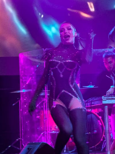 An image captured at the Mziuri amphitheater in Tsilkani, Georgia, showcases a female performer center-stage, bathed in vibrant purple and pink stage lighting. She is wearing a shimmering, dark, form-fitting costume with cutouts and intricate embellishments. Her mouth is open as if singing or shouting, and she is performing with energy, her left arm raised and her right hand appearing to touch her left hip. Behind her, a male DJ is visible, seated and operating DJ equipment, with a drum set partially obscured in the background. The overall environment suggests a live music performance or concert.