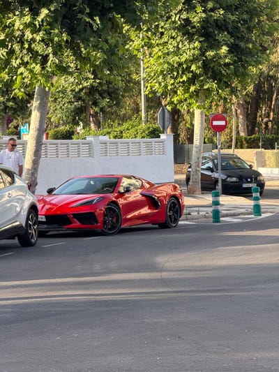 In Pla Parcial Platja, Spain, a red Chevrolet Corvette Stingray is parked on the side of a street, adjacent to a white SUV. The Corvette's sporty design and bright red color are prominent. A man wearing a white t-shirt and sunglasses stands near the white SUV, looking towards the Corvette. Behind the parked cars, a black Seat Ibiza is visible. The scene is lined with trees and a white, decorative wall, suggesting a residential or commercial area. Two green and white striped bollards stand on the sidewalk, indicating pedestrian crossing zones. A red circular sign with a white horizontal bar, signifying a 'no entry' or 'closed to all vehicles' restriction, is mounted on a pole nearby. The street is paved, and shadows from trees cast dappled patterns on the asphalt.