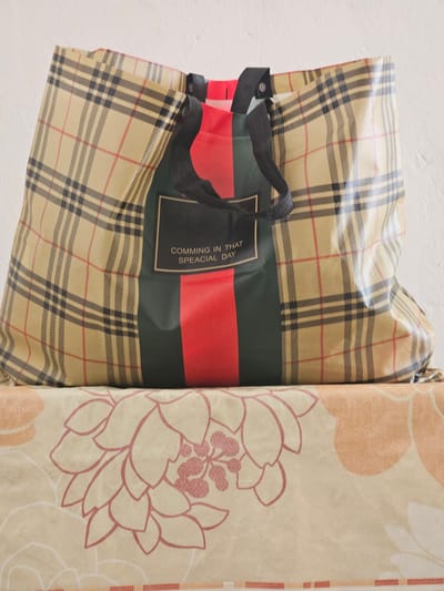A tan, plaid tote bag with black straps rests on a surface covered with a patterned fabric. The tote bag has a green panel with two vertical red stripes and a black rectangular label in the center. The label has white text that reads "COMMING IN THAT SPEACIAL DAY". The fabric underneath the bag features a beige background with large, stylized floral patterns in pink and orange. The scene is illuminated by soft lighting, casting subtle shadows. The background wall appears to be textured and white. This image is located in Saray, Azerbaijan.