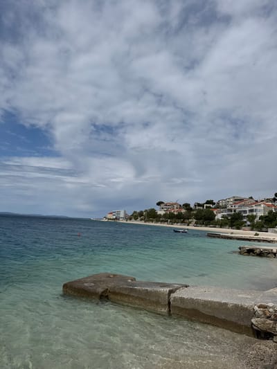 The image shows a view of the Adriatic Sea in Dugi Rat, Croatia. The camera is positioned on a concrete ramp that extends into the clear turquoise water. The water is shallow and the seabed is visible. In the distance, a shoreline lined with houses, trees, and a small boat is visible. The sky is cloudy with scattered white clouds against a blue backdrop. The image was likely taken in the afternoon.