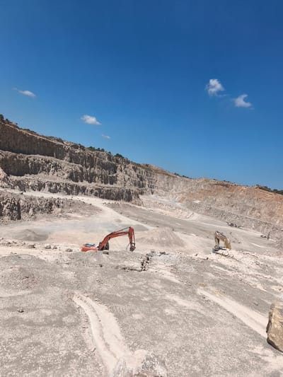 This is a photograph of an open-pit quarry in Riba-roja de Túria, Spain. The quarry exhibits large, stepped geological formations with visible layers of rock and soil. Two excavators are present: an orange one in the foreground and a yellow one further in the background. The sky is clear and blue with a few small white clouds. The ground is covered in loose rock, gravel, and sand. The scene appears to be one of active quarrying operations, with evidence of excavation and material removal.