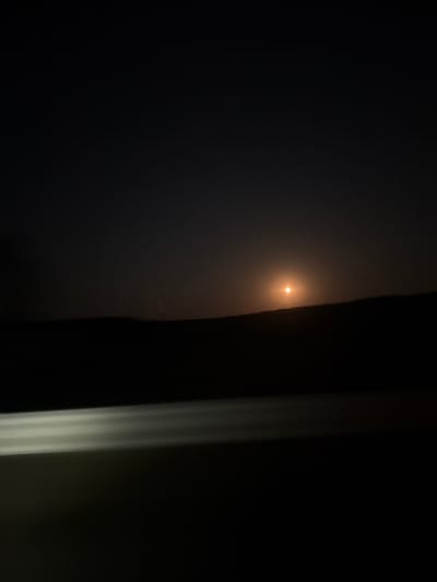 A nighttime scene features a bright, orange-tinted full moon illuminating a dark, silhouetted landscape. The moon is positioned slightly above the horizon, creating a soft glow that diffuses into the hazy sky. Below the moon, the dark undulating line of a hill or mountain range is visible. In the foreground, a horizontal band of bright, blurred white light streaks across the bottom half of the image, suggesting car headlights or a similar artificial light source. The sky is a deep, dark gradient with a few faint stars visible on the left. The overall impression is one of a rural or remote location at night, possibly viewed from a moving vehicle.