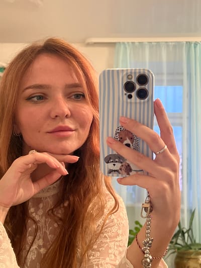 This is a selfie taken in portrait orientation. The subject is a young woman with long, reddish-blonde hair. She is holding a smartphone with a light blue and white striped case, decorated with cartoonish dog images, in her right hand, pointing the camera towards a mirror. Her left hand is resting gently on her cheek, and she is wearing two rings, one on her index finger and one on her ring finger. Her eyes are green, and she is wearing makeup, including eyeliner.
The background appears to be an indoor setting, possibly a room with a window covered by light blue blinds. The lighting suggests daytime. The woman is wearing a white, lacy garment, which could be a dress or a top. The overall mood conveyed by the image is relaxed and casual. There is no visible text in the image. The metadata indicates the location is Mezhdurechenskiy, Russia.