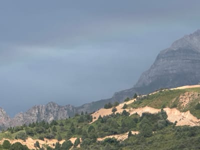 A view of a mountainous landscape under a cloudy sky, with a hint of a rainbow visible in the distance. The foreground features a green, treed hillside with exposed sandy soil, indicative of a man-made excavation or landslide. In the background, rugged mountain peaks rise, some with visible rock formations. The scene suggests a natural environment with signs of human intervention or geological activity.