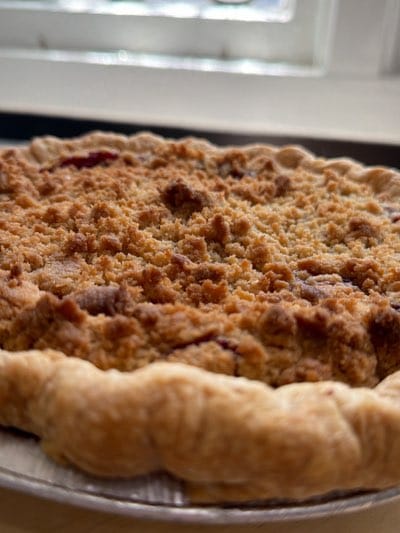 The image shows a close-up of a freshly baked crumble-topped fruit pie with a golden-brown crust. The pie is sitting on what appears to be a metal baking sheet. The topping is textured with small, irregular crumbs. There is a bright, blurred window in the background. It is impossible to determine the location based solely on the image.