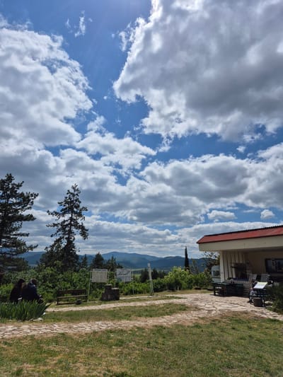 The image depicts an outdoor scene in Raianovtsi, Bulgaria. The foreground features trees and vegetation. A partially visible building with a red roofline is present on the right side. A stone path extends from the building into the grassy area. Mountains are visible in the distance. The sky is filled with white, billowing clouds against a blue backdrop. A signpost and bench are located by the path. Two people are seated near the trees.