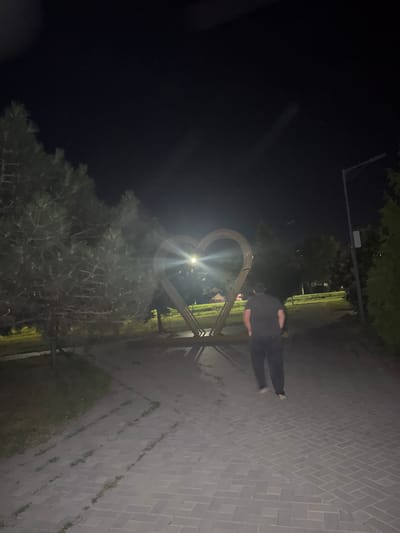 A person walks away from the camera at night in a park in Egorlykskaya, Russia. The person is wearing a black t-shirt and black pants. In the background, there is a large heart-shaped sculpture illuminated by a bright light. The sculpture is made of metal. The ground is paved with light-colored bricks. Trees and grass are visible on either side of the path. A lamppost is also visible on the right side of the image. The sky is dark.