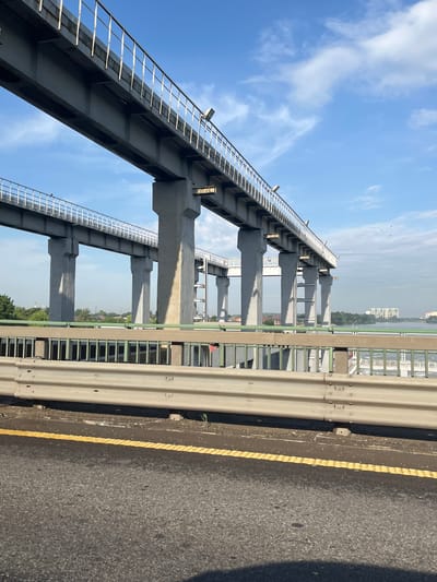 The image captures an elevated concrete bridge structure under a partly cloudy blue sky. The bridge, supported by numerous large concrete pillars, extends horizontally across the frame. A secondary, lower bridge runs parallel to the first, also supported by pillars. Both bridges feature railings along their edges. In the foreground, a road with a yellow dividing line is visible, and a metal guardrail separates the road from the bridge structure. Beyond the bridge, a body of water can be seen, with buildings and trees along its banks. The scene appears to be an urban or suburban landscape, possibly in Tlustenkhabl, Russia, given the context of the prompt. There are no visible people or animals in the image, and the overall atmosphere is one of infrastructure and a somewhat overcast day.