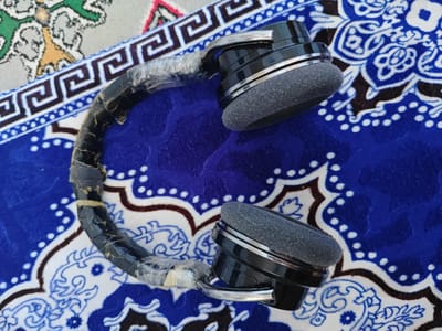 A pair of black headphones with silver accents are positioned on a blue and white patterned rug. The headphones appear worn, with the headband covered in clear tape and dark tape. The earcups are grey and circular. The rug has a central floral design with white and blue elements. In the background, a portion of another rug or fabric with green, white, and yellow patterns is visible. The lighting suggests an indoor setting. The image is taken from a top-down perspective.