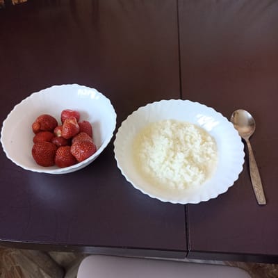 An overhead shot shows two white, fluted bowls and a spoon on a dark brown table. The bowl on the left contains about ten strawberries. The bowl on the right contains rice pudding. A spoon rests on the table to the right of the bowl of rice pudding. The table is dark brown and appears to be a wooden surface. The floor visible at the bottom of the image is tiled and a light gray chair is partially visible. The image is a photograph of breakfast food. This scene does not depict Shakhunya, Russia.